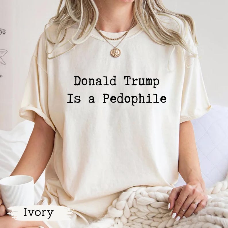 以下が含まれることがあります： 「Donald Trump Is a Pedophile」の黒い文字がプリントされたアイボリー色のTシャツ。クルーネックのシャツです。「Ivory」の文字が画像の下部に印刷されています。