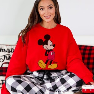 Op de afbeelding: Een rode sweatshirt met een Mickey Mouse-afbeelding. De Disney-sweatshirt heeft lange mouwen en wordt gecombineerd met een zwart-wit geruite pyjamabroek. De afbeelding toont ook een kussen met de tekst "Christ Tre" en een rood-witte mok.
