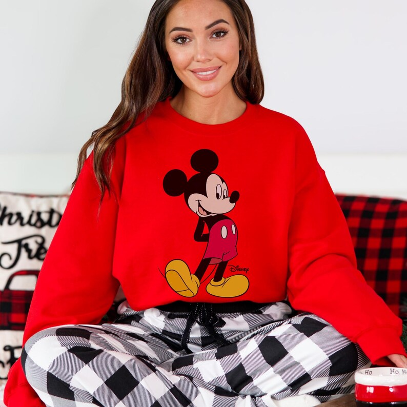 Op de afbeelding: Een rode sweatshirt met een Mickey Mouse-afbeelding. De Disney-sweatshirt heeft lange mouwen en wordt gecombineerd met een zwart-wit geruite pyjamabroek. De afbeelding toont ook een kussen met de tekst "Christ Tre" en een rood-witte mok.