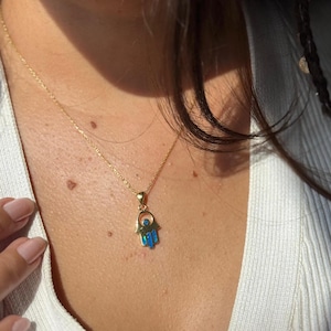 Könnte beinhalten: Eine goldfarbene Halskette mit einem Hamsa-Hand-Anhänger. Der Anhänger hat blaue und türkisfarbene Emaille-Details. Die Halskette wird von einer Person getragen, die einen weißen Rippenpullover trägt.
