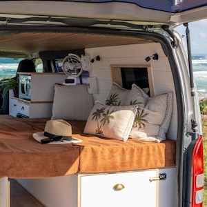Puede incluir: Vista interior de una furgoneta camperizada con cama, almohadas y microondas. La cama tiene una funda de pana marrón y las almohadas diseños de palmeras. Un sombrero de paja y gafas de sol descansan sobre la cama. La furgoneta está cerca del mar.