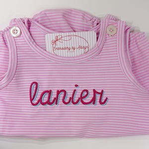 May include: Pink and white striped baby romper with the name "lanier" embroidered in red with a blue shadow. The romper has button closures on the shoulders and a tag that reads "Embroidery by Adisyn".
