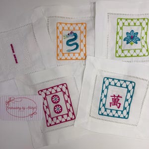 K&ouml;nnte beinhalten: Wei&szlig;e, bestickte Leinenuntersetzer mit verschiedenen Designs. Ein Untersetzer zeigt einen blauen Drachen, ein anderer eine Lotusblume, und andere geometrische Muster und chinesische Schriftzeichen. Die Untersetzer sind quadratisch mit dekorativen N&auml;hten.