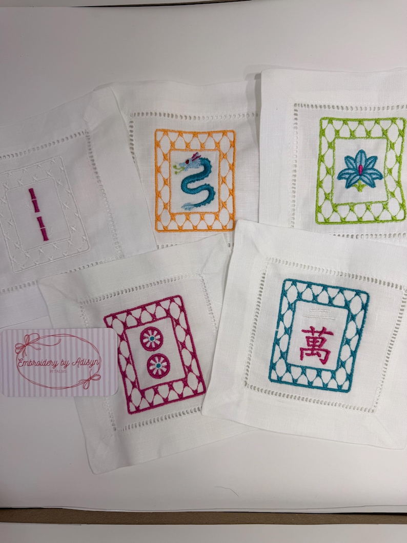 K&ouml;nnte beinhalten: Wei&szlig;e, bestickte Leinenuntersetzer mit verschiedenen Designs. Ein Untersetzer zeigt einen blauen Drachen, ein anderer eine Lotusblume, und andere geometrische Muster und chinesische Schriftzeichen. Die Untersetzer sind quadratisch mit dekorativen N&auml;hten.