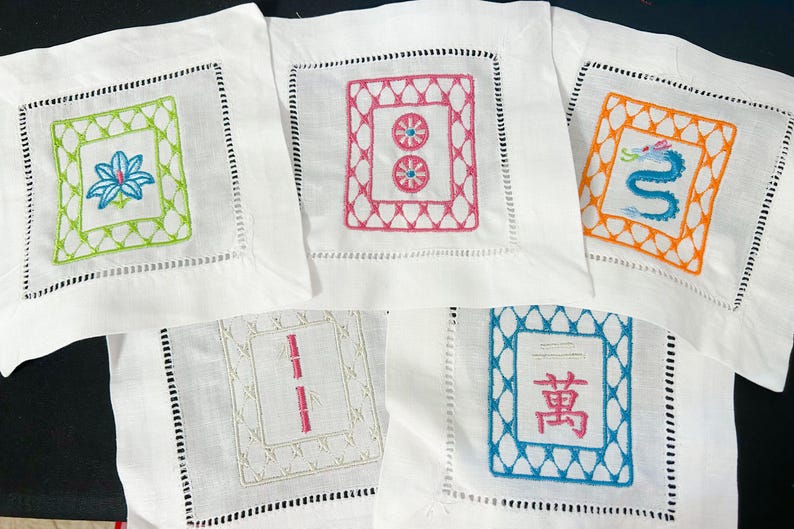 K&ouml;nnte beinhalten: F&uuml;nf wei&szlig;e Leinenuntersetzer mit gestickten Designs. Jeder Untersetzer hat ein anderes farbenfrohes Design in einem quadratischen Rahmen. Zu den Designs geh&ouml;ren eine Blume, zwei Kreise, ein Drache, Bambusst&auml;be und ein chinesisches Schriftzeichen.