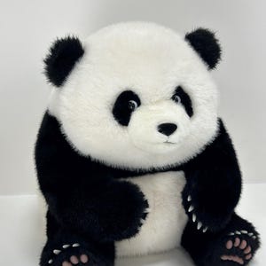May include: A plush panda bear with black and white fur. The panda has black ears, eyes, and limbs, with a white face and body. The bear is sitting upright with its paws visible, and has a sweet expression.