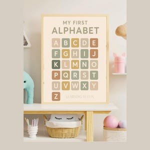Op de afbeelding: Een ingelijste alfabetposter met de tekst "MY FIRST ALPHABET" en de letters A-Z in een raster. De letters zijn in verschillende pastelkleuren. De tekst "LEARNING IS FUN" staat onderaan. De poster staat op een witte plank met decoratieve items.