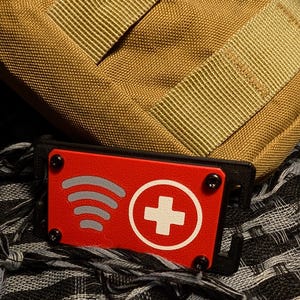 Pode incluir: Um patch retangular vermelho com uma cruz branca e três linhas curvas, fixado com parafusos pretos. O patch está sobre uma base preta, contra um fundo de tecido bege e um tecido estampado em preto e branco.