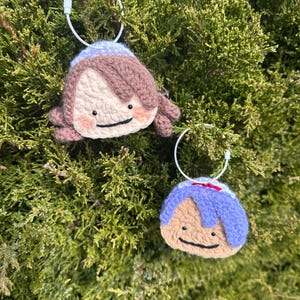 May include: Two handmade crochet doll keychains. One keychain features a doll with brown hair and a light blue hat. The other keychain has blue hair and a blue hat. Both dolls have a smiling face and a white metal ring.