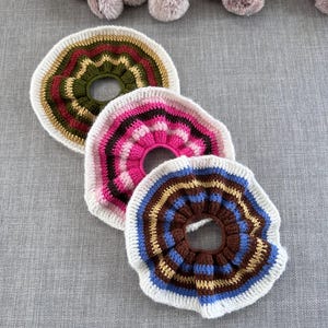 Op de afbeelding: Drie handgemaakte gehaakte onderzetters in verschillende kleuren. Eén onderzetter heeft olijfgroen, rood en goud, een andere roze en wit, en de derde bruin, blauw en goud. Elke onderzetter heeft een witte rand en een centraal gat.
