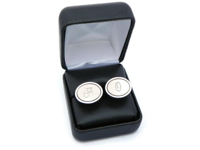 May include: Silver cufflinks with the letters "A" and "O" engraved on them, presented in a black leatherette gift box with a white satin lining. The cufflinks have an oval shape and a polished finish.