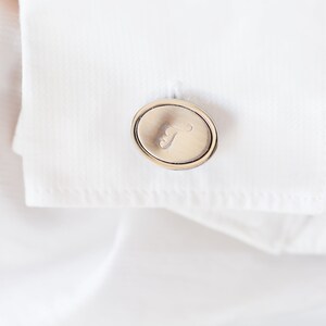 May include: Close-up of a silver cufflink with an oval design and the letter "T" engraved on it. The cufflink is attached to a white dress shirt, creating a classic and elegant look. The shirt's fabric is crisp and smooth.