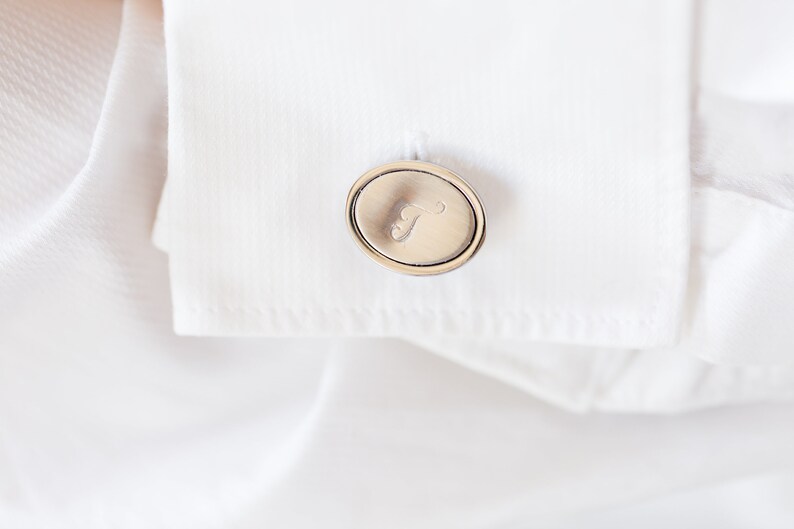 May include: Close-up of a silver cufflink with an oval design and the letter "T" engraved on it. The cufflink is attached to a white dress shirt, creating a classic and elegant look. The shirt's fabric is crisp and smooth.