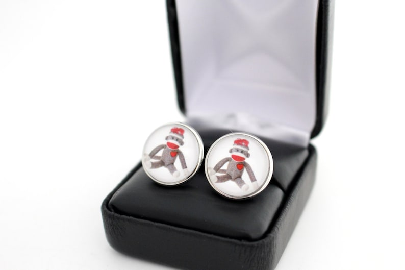 May include: A pair of silver cufflinks with a round, clear glass design featuring a cartoon sock monkey wearing a red heart. The cufflinks are in a black velvet box.