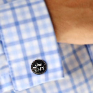 May include: A close-up of a black cufflink with the word "Dad" and the date "9/24/78" in white script. The cufflink is on a blue and white checkered shirt.