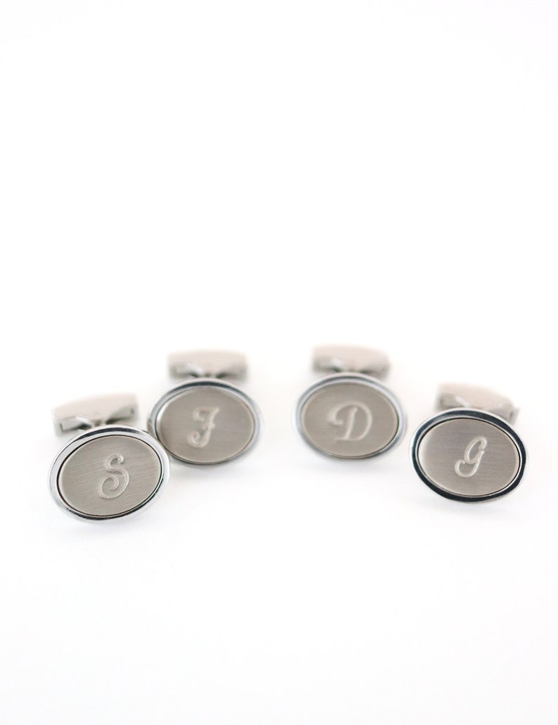 May include: Silver-toned oval cufflinks with engraved initials. The cufflinks have a brushed metal finish and a polished silver border. The initials "S", "F", "D", and "g" are displayed on the face of each cufflink. The cufflinks are presented on a white background.