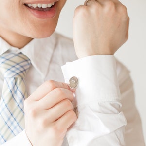 May include: A close-up of a white dress shirt with a light blue and white plaid tie. The person is fastening a silver cufflink on the shirt sleeve. The cufflink is round with a silver border.