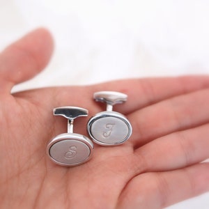 May include: Two silver cufflinks with oval faces, each engraved with a single letter. One cufflink has the letter "S" and the other has the letter "J". The cufflinks are displayed on a light background.