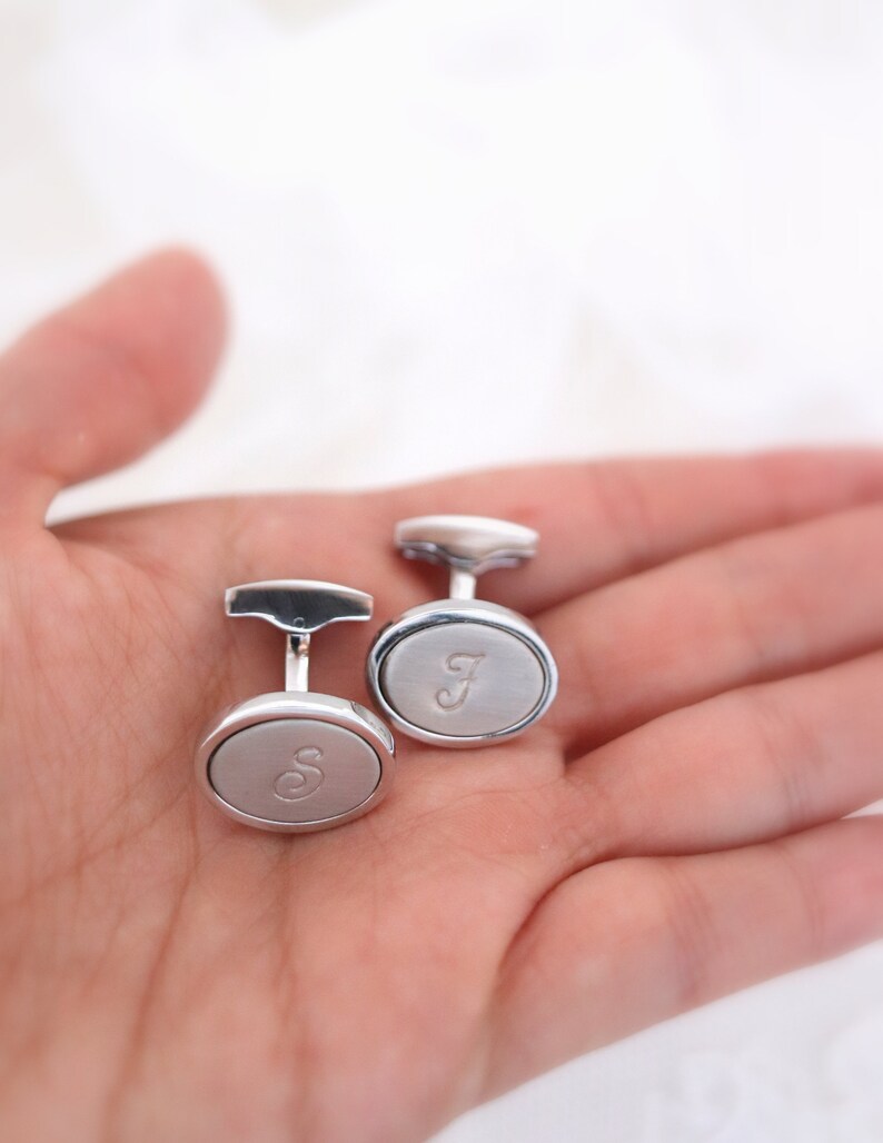 May include: Two silver cufflinks with oval faces, each engraved with a single letter. One cufflink has the letter "S" and the other has the letter "J". The cufflinks are displayed on a light background.