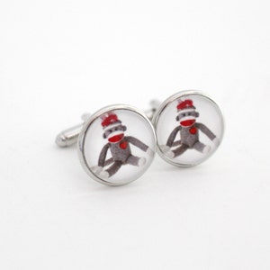 May include: A pair of silver cufflinks with a round, clear glass design featuring a cartoon sock monkey wearing a red hat.