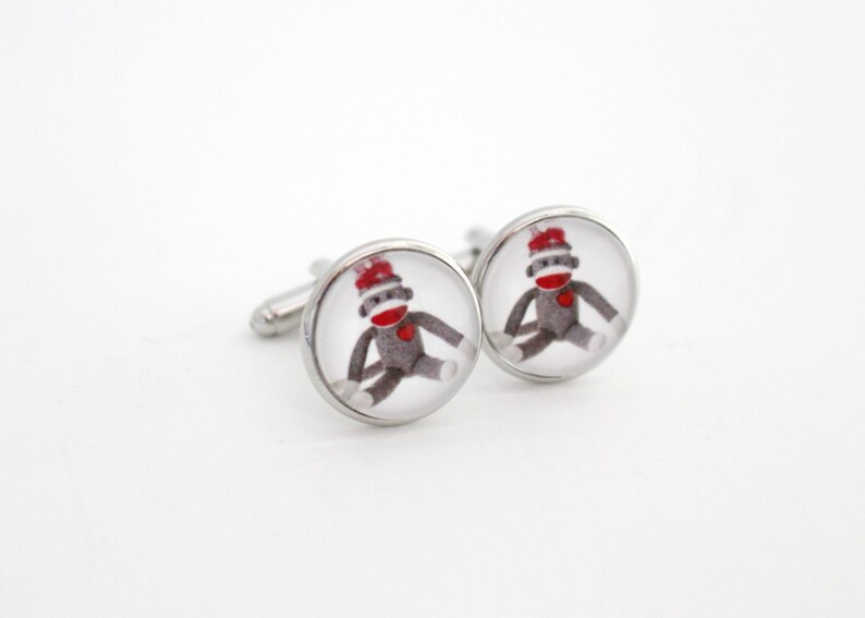 May include: A pair of silver cufflinks with a round, clear glass design featuring a cartoon sock monkey wearing a red hat.