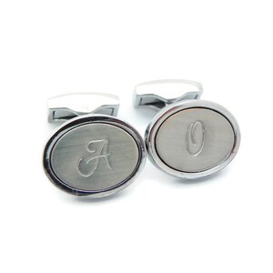 May include: A pair of silver cufflinks with an oval face. One cufflink features the letter "A" and the other has the letter "O" engraved on the face. The cufflinks have a polished finish and a rectangular post.