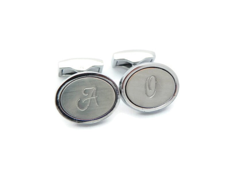 May include: A pair of silver cufflinks with an oval face. One cufflink features the letter "A" and the other has the letter "O" engraved on the face. The cufflinks have a polished finish and a rectangular post.
