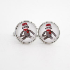 May include: A pair of silver cufflinks with a round, clear glass design featuring a sock monkey wearing a red hat.