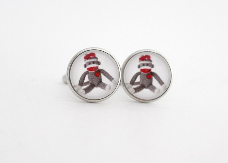 May include: A pair of silver cufflinks with a round, clear glass design featuring a sock monkey wearing a red hat.