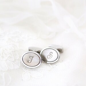 May include: Silver-toned oval cufflinks with engraved initials "J" and "S". The cufflinks have a brushed metal finish and a polished silver border. They are displayed on a white, textured surface.