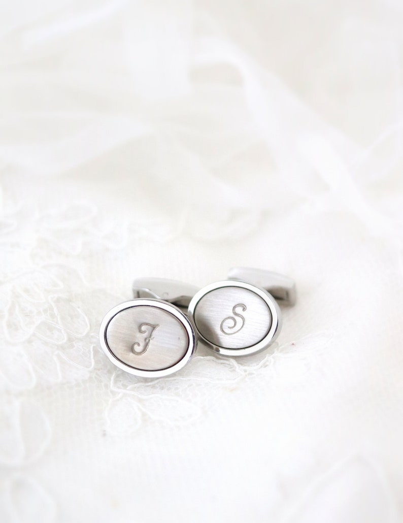 May include: Silver-toned oval cufflinks with engraved initials "J" and "S". The cufflinks have a brushed metal finish and a polished silver border. They are displayed on a white, textured surface.