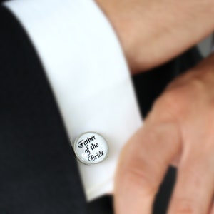 May include: A silver cufflink with the text "Father of the Bride" in black script. The cufflink is on a white dress shirt cuff, with a black suit jacket in the background.