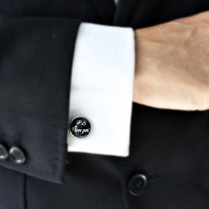 May include: A black and silver cufflink with the text "P.S. I love you" in white script. The cufflink is on a white dress shirt cuff, paired with a black suit jacket. The image is a close-up of the cufflink and shirt.