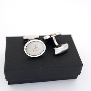 May include: Silver cufflinks with an oval face and the letter "S" engraved on a brushed metal surface. The cufflinks are presented on a black box, set against a white background. A classic accessory.