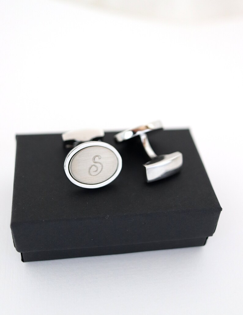 May include: Silver cufflinks with an oval face and the letter "S" engraved on a brushed metal surface. The cufflinks are presented on a black box, set against a white background. A classic accessory.