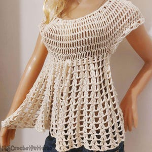 Puede incluir: Top de crochet color crema con cuello redondo y mangas cortas. El top presenta un patrón de puntada abierto y texturizado y un dobladillo ligeramente acampanado. La prenda se muestra en un maniquí.