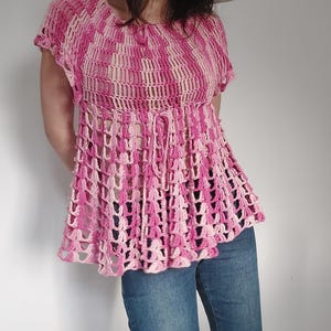 May include: A pink and white crochet top with short sleeves and a flared hem. The top features a lattice-like design and a drawstring at the waist. The model is wearing blue jeans and a sun hat. The top is handmade.