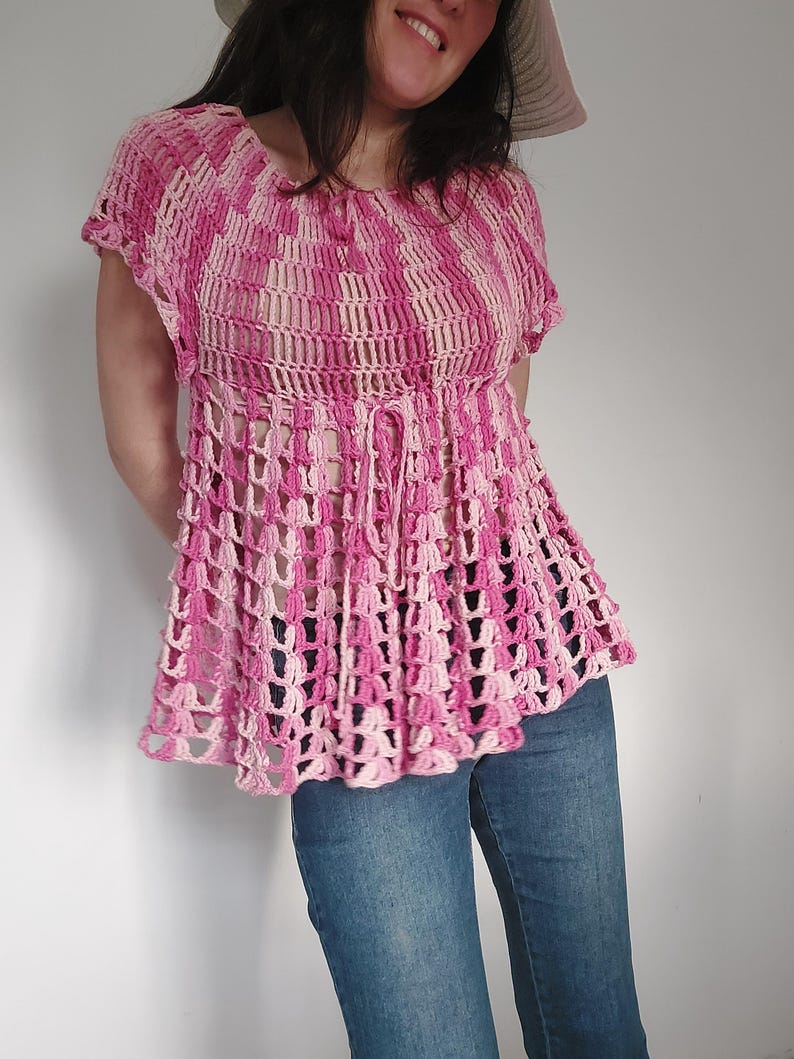 May include: A pink and white crochet top with short sleeves and a flared hem. The top features a lattice-like design and a drawstring at the waist. The model is wearing blue jeans and a sun hat. The top is handmade.