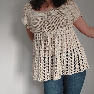 May include: Cream-colored crochet top with a loose, open weave design. The top features short sleeves and a drawstring detail at the neckline. The bottom of the top flares out slightly. The top is paired with blue jeans.