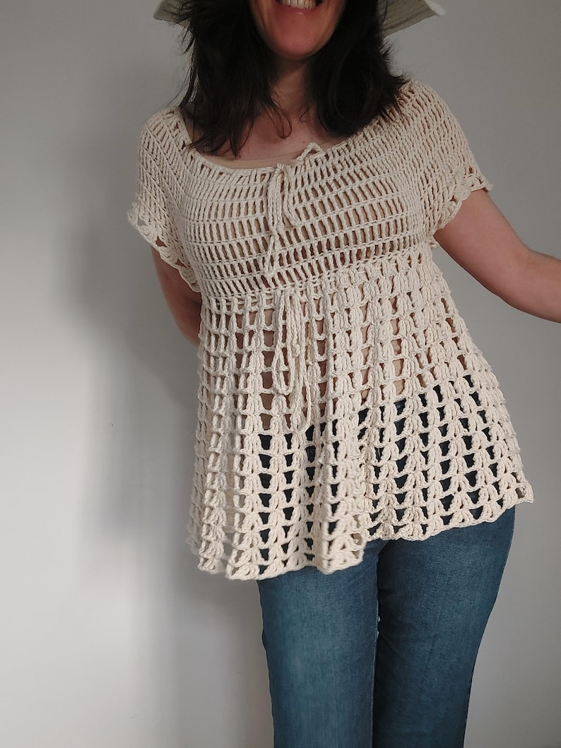 May include: Cream-colored crochet top with a loose, open weave design. The top features short sleeves and a drawstring detail at the neckline. The bottom of the top flares out slightly. The top is paired with blue jeans.