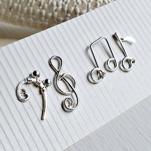 May include: Four silver wire music note charms. The charms are a treble clef, a quarter note, an eighth note, and a whole note. The charms are arranged in a row on a white background.