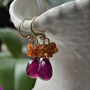 May include: Gold-tone earrings with a cluster of orange beads and two teardrop-shaped, deep pink gemstones. The earrings are displayed against a blurred background with greenery and a white ceramic vase.