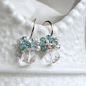 May include: A pair of gold-tone earrings featuring clusters of small, light blue and white beads, with a clear, faceted crystal pendant. The earrings are displayed against a white background.
