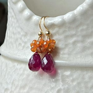 May include: Gold-tone earrings with a cluster of orange beads and two faceted, teardrop-shaped, deep red gemstones. The earrings are hanging on a white ceramic vase. The earrings are a jewelry item.