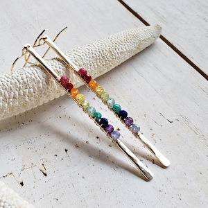 May include: A pair of gold-colored earrings with a linear design. Each earring features a row of colorful beads in a rainbow spectrum, attached to a long, rectangular gold bar. The beads include red, orange, yellow, green, blue, and purple.