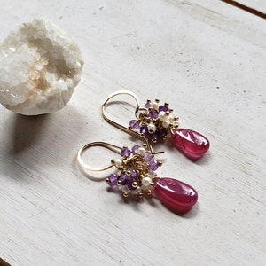 May include: Gold earrings with a cluster of purple and white gemstones and a teardrop-shaped red gemstone dangling from each earring.