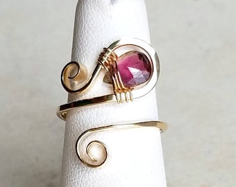 May include: Gold wire wrapped ring with a pink and clear gemstone. The ring is on a white display stand.