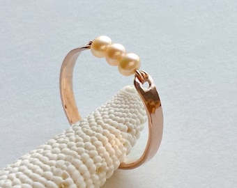 May include: A delicate rose gold ring with three small, off-white pearls set in a row. The ring is on a white, textured surface.