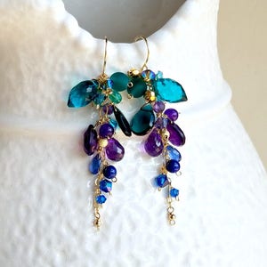 May include: Gold dangle earrings with teal, purple, and blue gemstone beads. The earrings feature a cluster of faceted teal gemstones at the top, followed by a cascade of purple and blue beads.