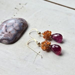 May include: Gold-tone earrings with a cluster of orange beads and a teardrop-shaped, deep pink gemstone. A polished, oval-shaped stone with swirling patterns in shades of gray, brown, and white is also present.